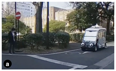 図a：実験で実験参加者に提示された映像の例。実験参加者は手前から車両を見ている。道路の向こう側に別の歩行者が立っている。(a)車両の視線が実験参加者を向いている（停止する場合）